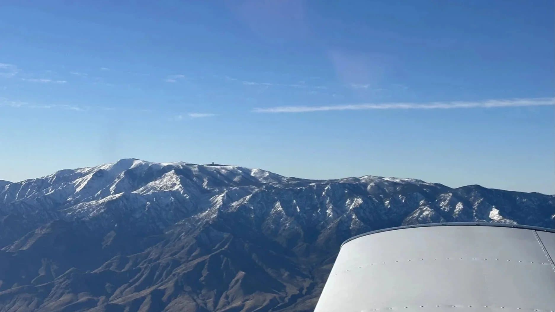 Snow-capped mountain ridge seen beyond a training aircraft's wing under a clear blue sky